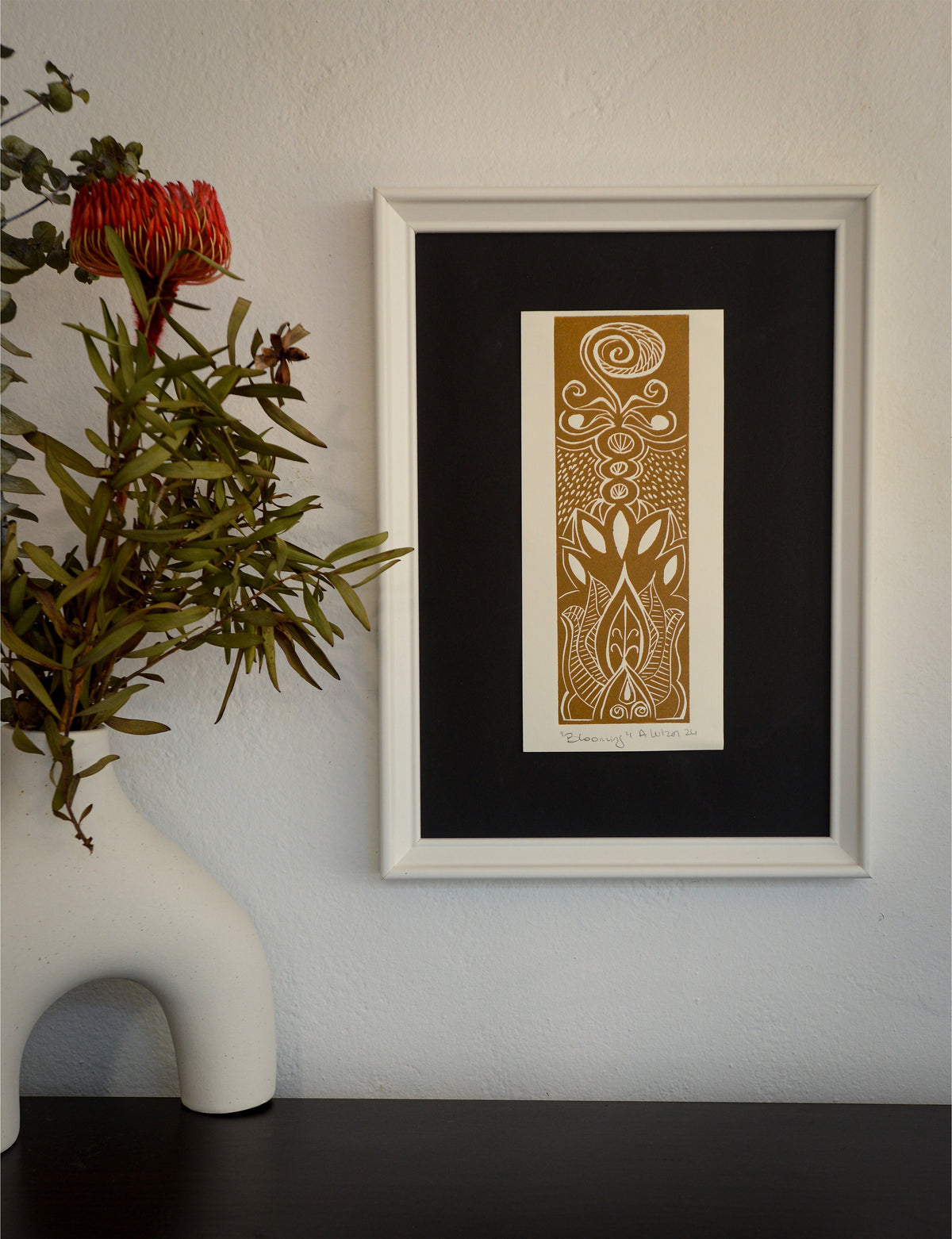 The Blooming linocut in a black frame with a wide mount, beside a vase