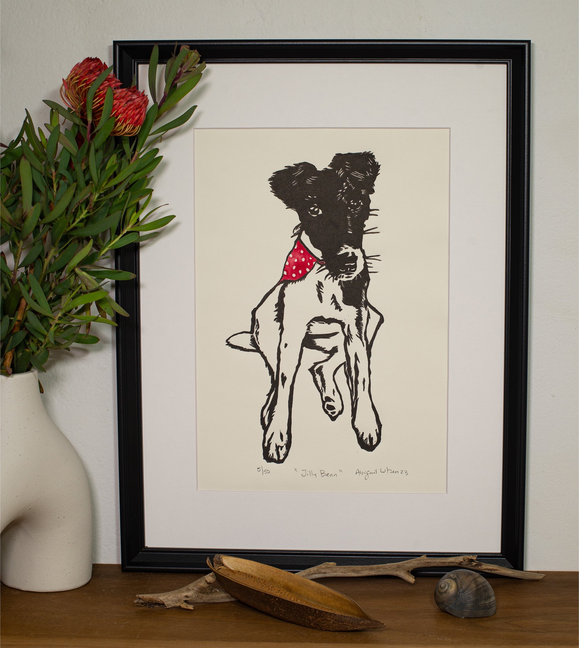 The Jilly Bean linocut in a white frame, beside a vase of native foliage
