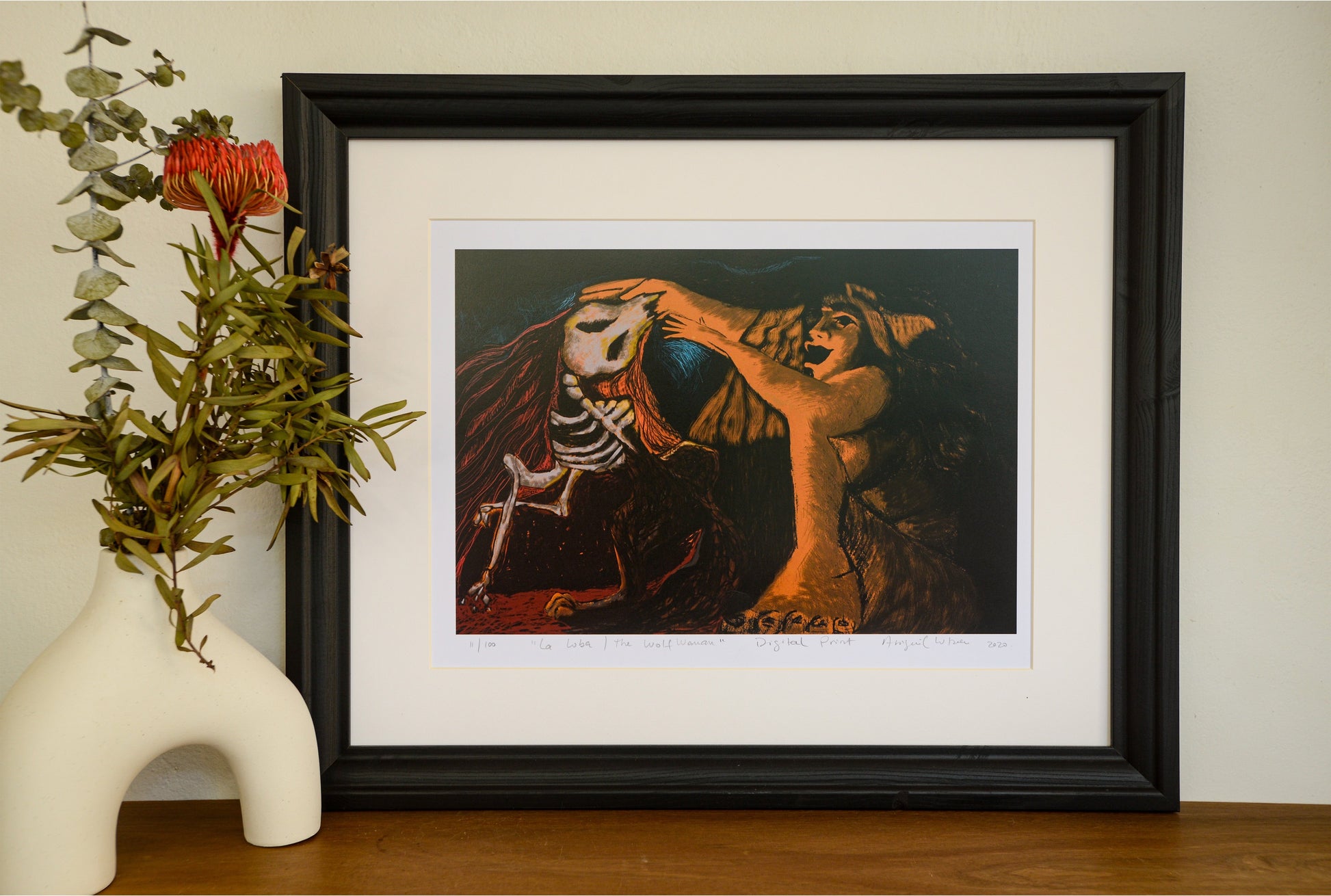 The La Loba linocut in a black frame with a wide mount, beside a vase of native flowers
