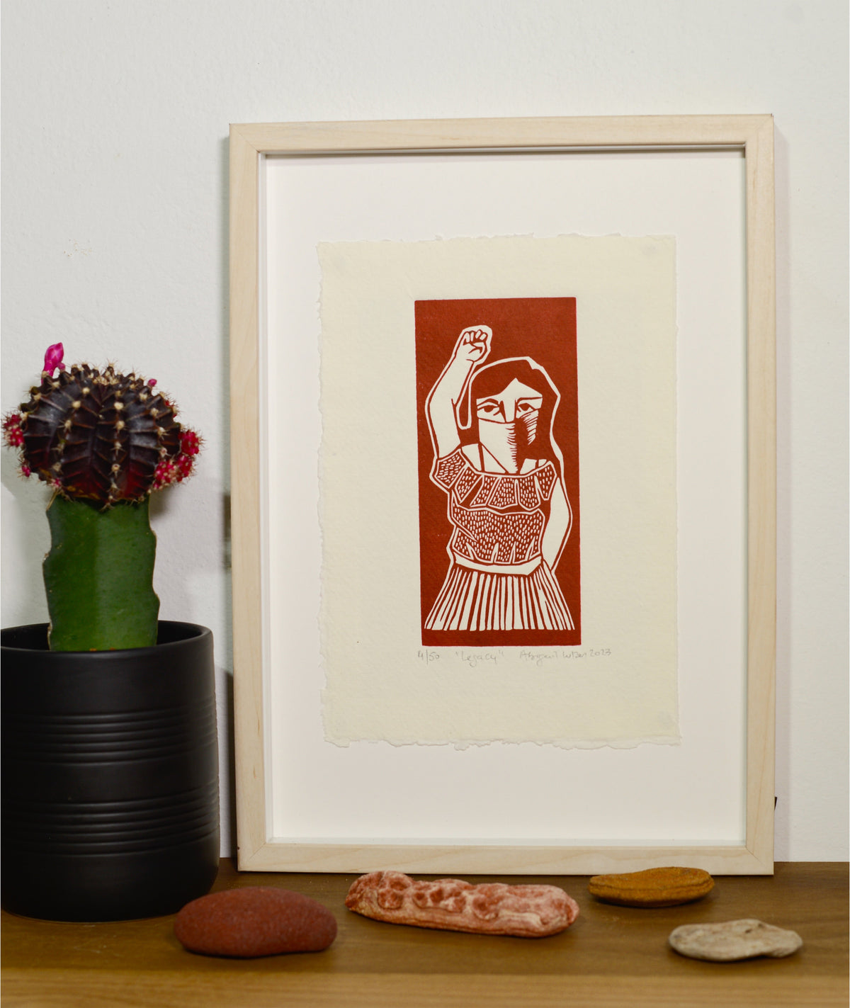 The Legacy linocut in a white frame, beside a potted plant