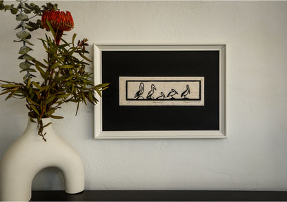 The Pelicans linocut in a long black frame, hung beside a vase of native flowers