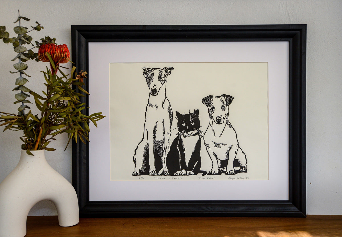 The Rocky, Sootie and Bluebelle linocut in a black frame, beside a vase of native flowers