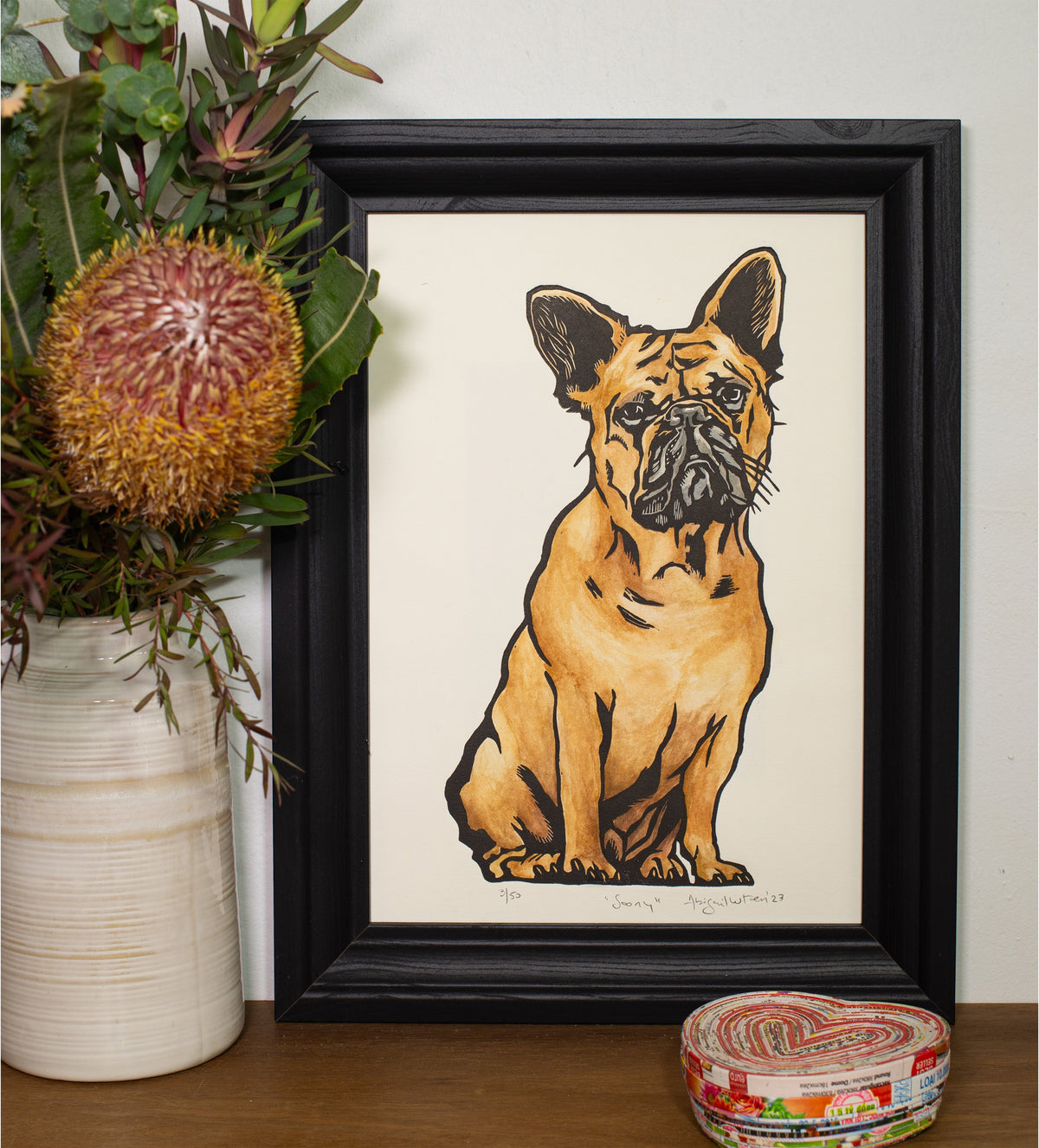 The Soony linocut in a black frame, beside a vase of native flowers