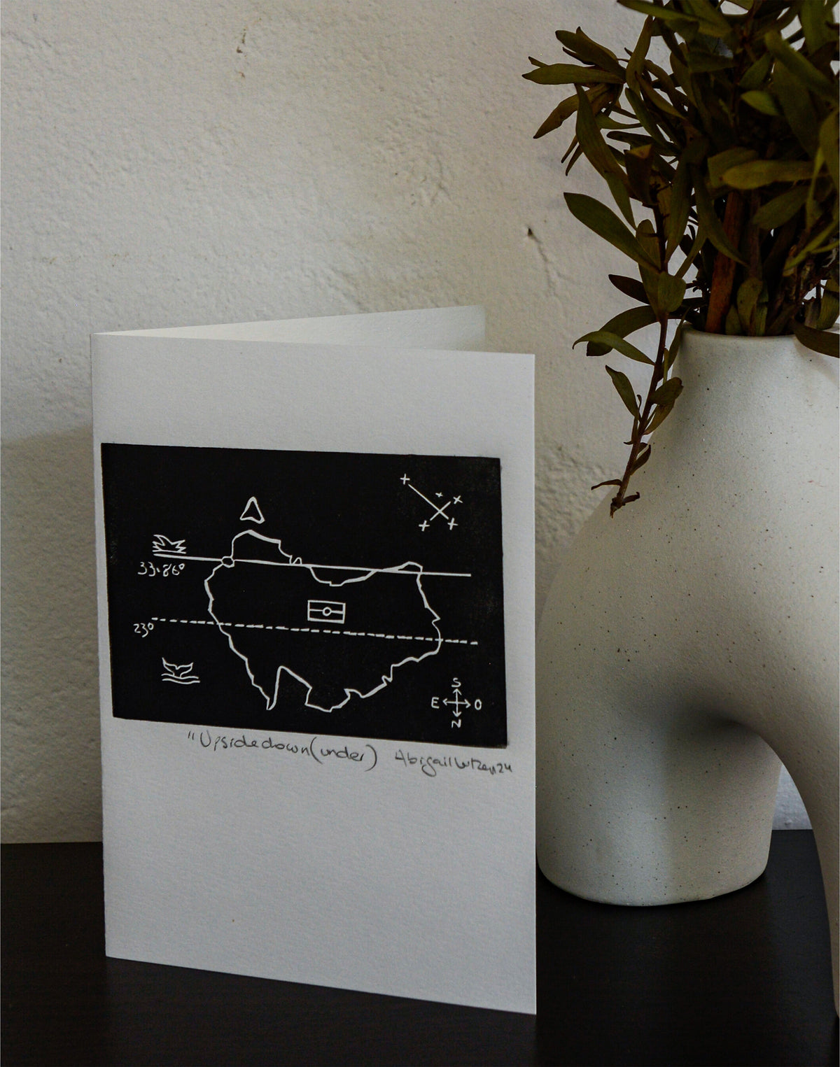 The Upside Down(under) linocut postcard standing upright beside a vase