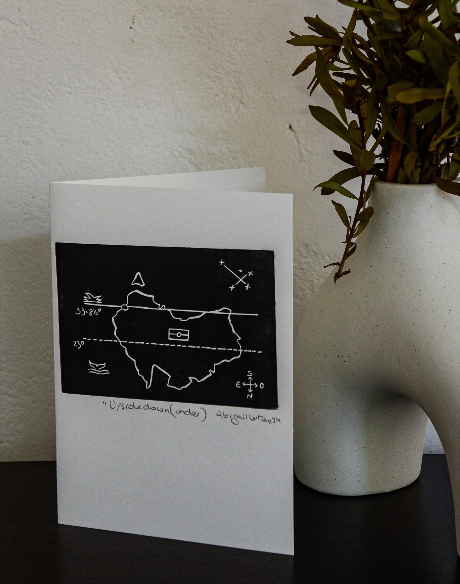 The Upside Down(under) linocut postcard standing upright beside a vase